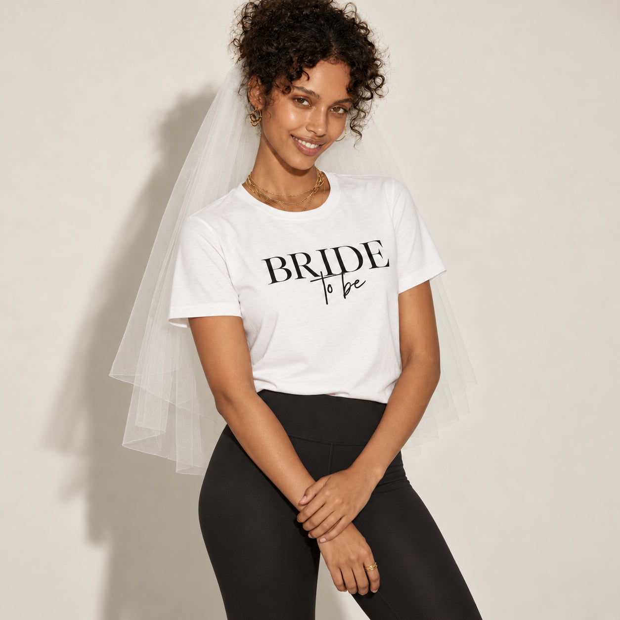 A woman wearing a white graphic t-shirt with 'BRIDE to be' printed in black, paired with black leggings and a sheer white bridal veil, posing against a light beige wall.