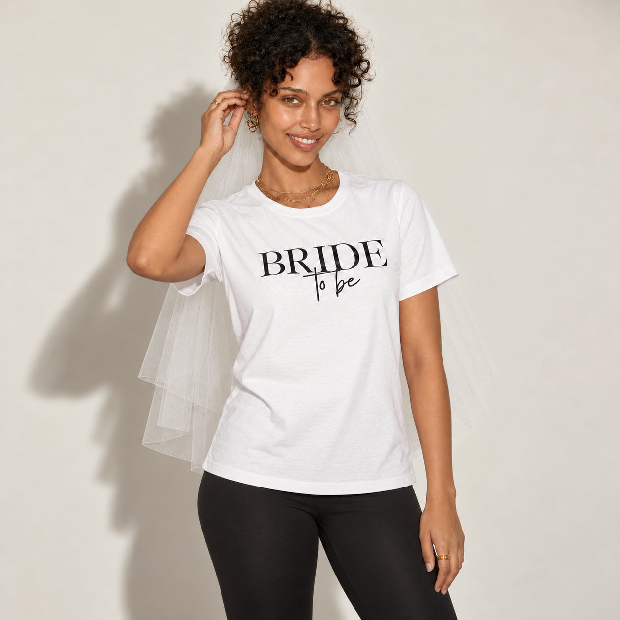 A woman wearing a white graphic t-shirt with 'BRIDE to be' printed in black, styled with a white tulle bridal veil and black leggings against a light beige background.