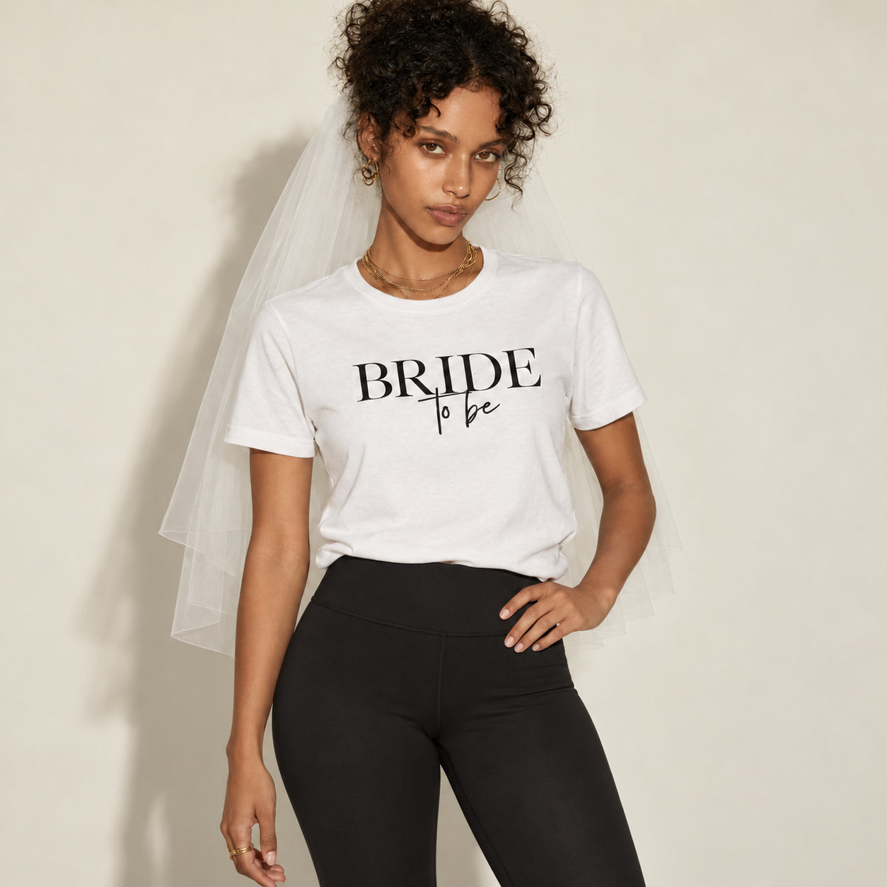 A woman wearing a white crewneck t-shirt with 'BRIDE to be' printed in black text, styled with a white tulle bridal veil and black leggings, posing against a neutral beige background.