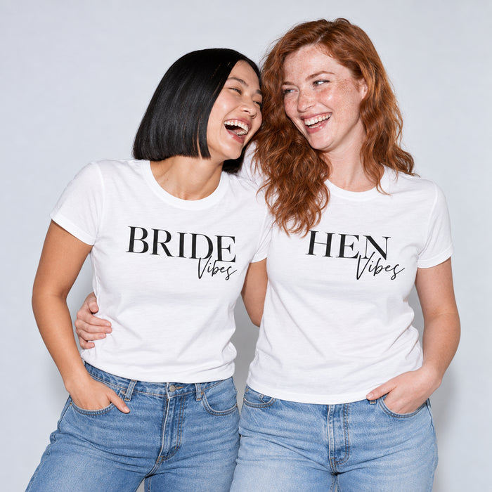 Two women wearing matching white graphic t-shirts that read 'BRIDE Vibes' and 'HEN Vibes' in black lettering, paired with blue jeans.