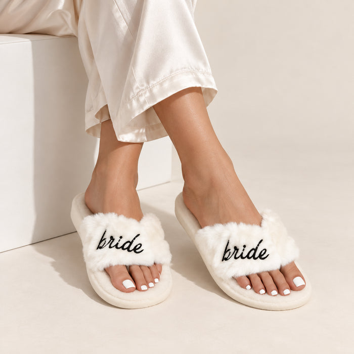 A person wearing fluffy white slide slippers with the word 'bride' embroidered in black script across the straps, paired with cream satin pants. 
