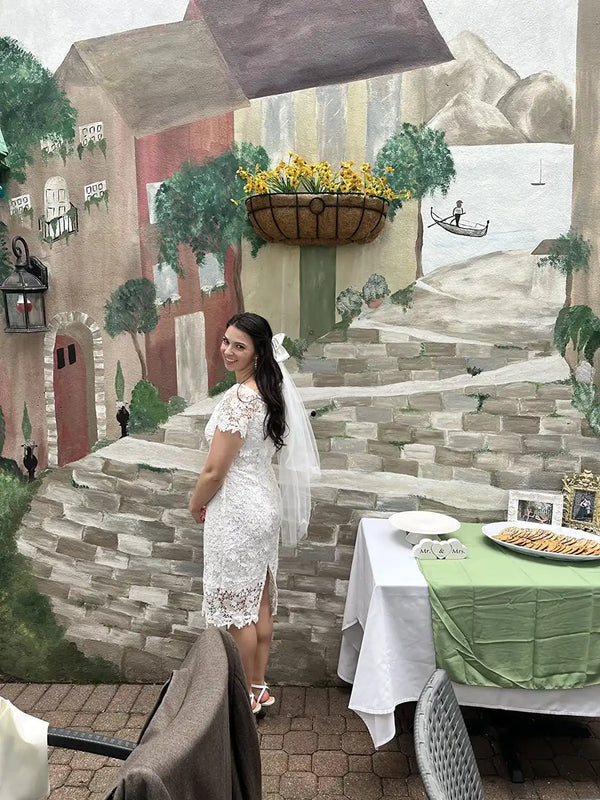 Bride to be in a white dress standing in front of a scenic mural with a table set for a meal.