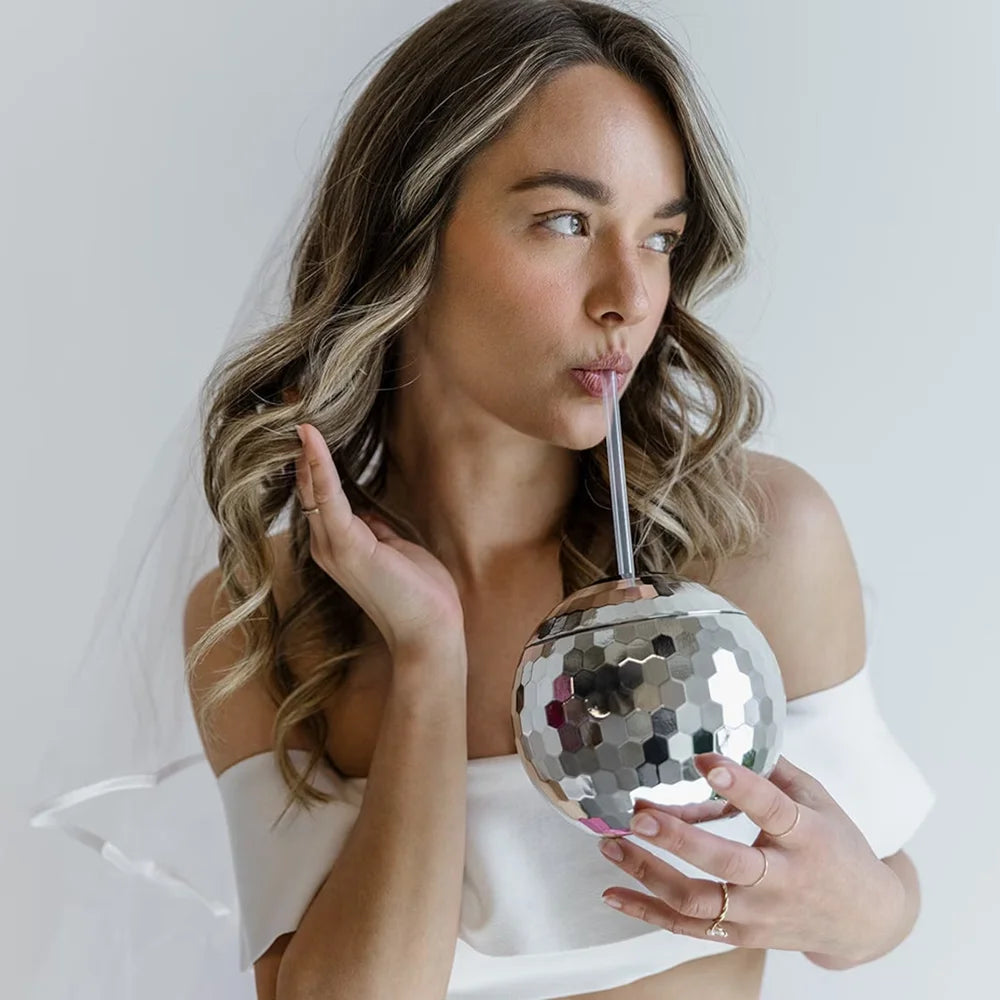 Woman drinking from a silver disco ball with a straw against a plain background