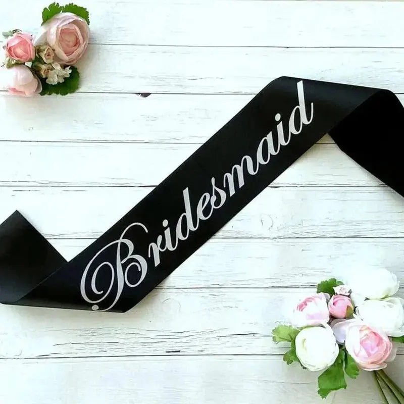 Black sash with 'Bridesmaid' text on a white wooden surface with floral decorations.