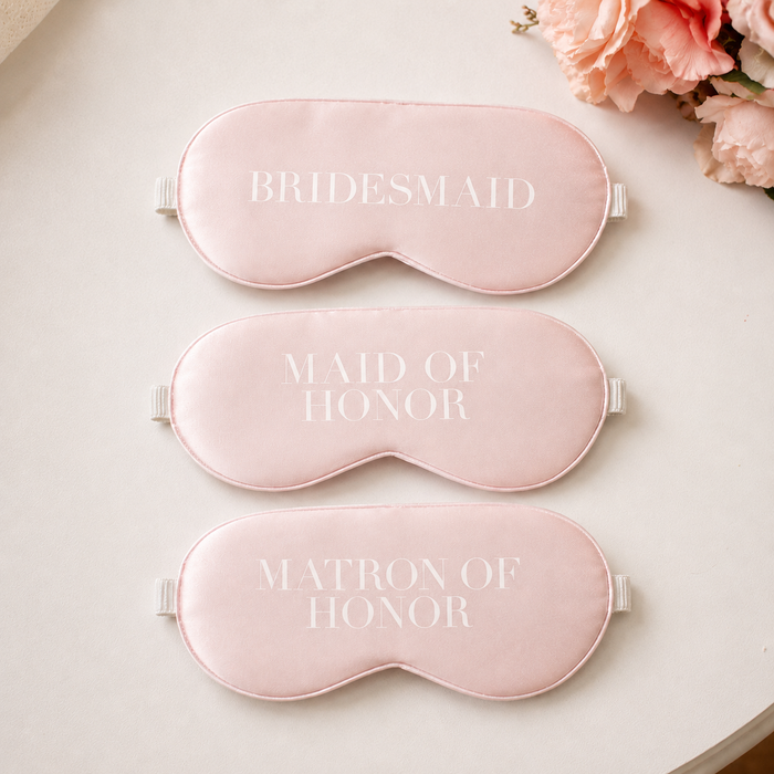 Blush pink satin sleep masks for Bridesmaid, Maid of Honor and Matron of Honor displayed on a light neutral tabletop with soft peach floral accents.