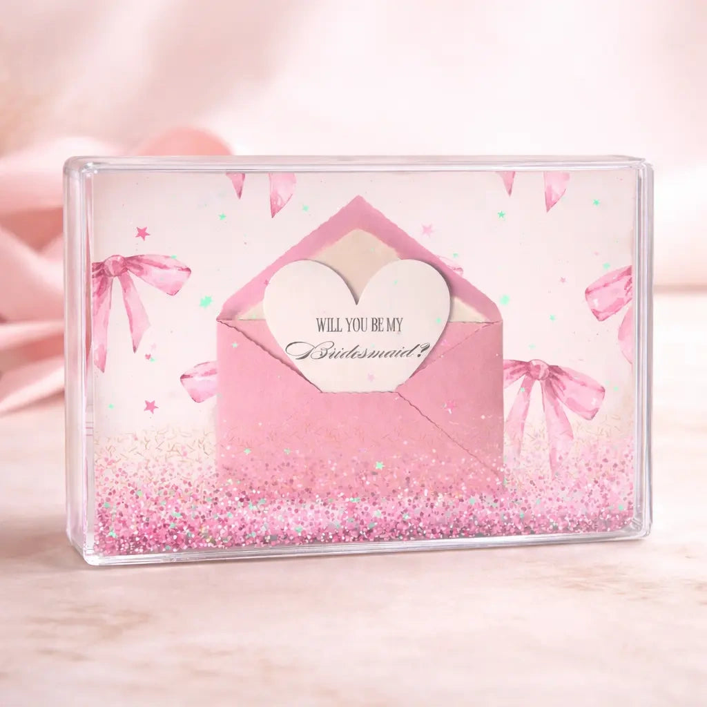 Close-up of a bridesmaid proposal confetti block with a pink envelope and heart-shaped “Will you be my Bridesmaid?” card inside, finished with pink glitter and iridescent confetti details.