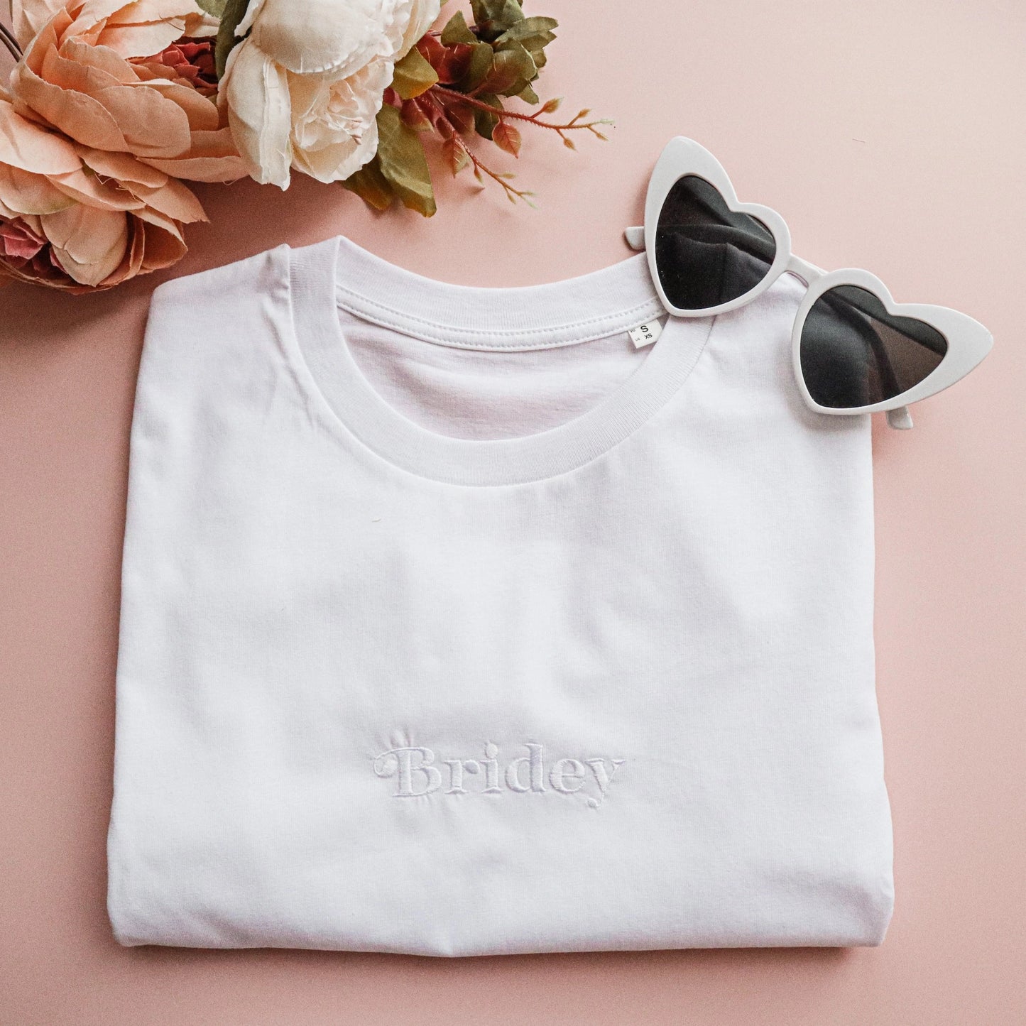 Folded white t-shirt with 'Bridey' embroidery on a pink surface with heart-shaped sunglasses and flowers.