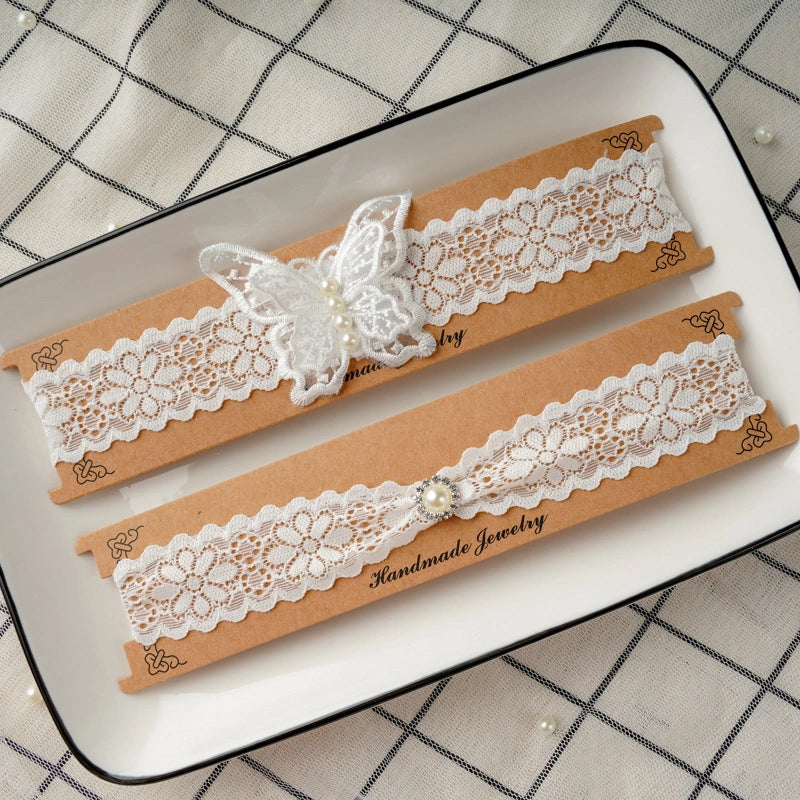 Two white lace bridal garters with butterfly and pearl embellishments, displayed on brown kraft paper bands inside a white rectangular tray with a black rim.