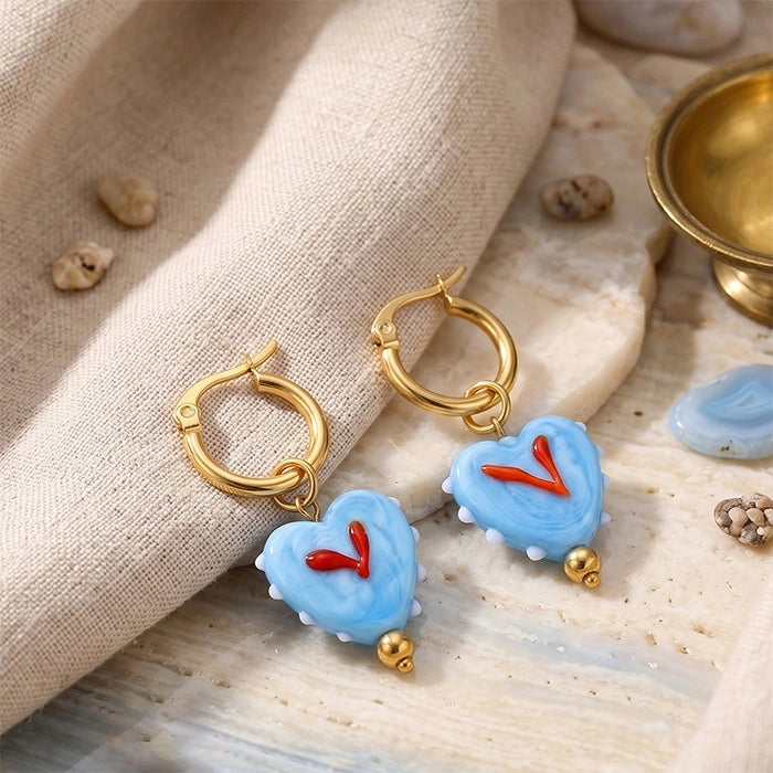 A pair of gold hoop earrings with light blue heart-shaped charms and small red heart details, styled on a beige fabric and stone surface.