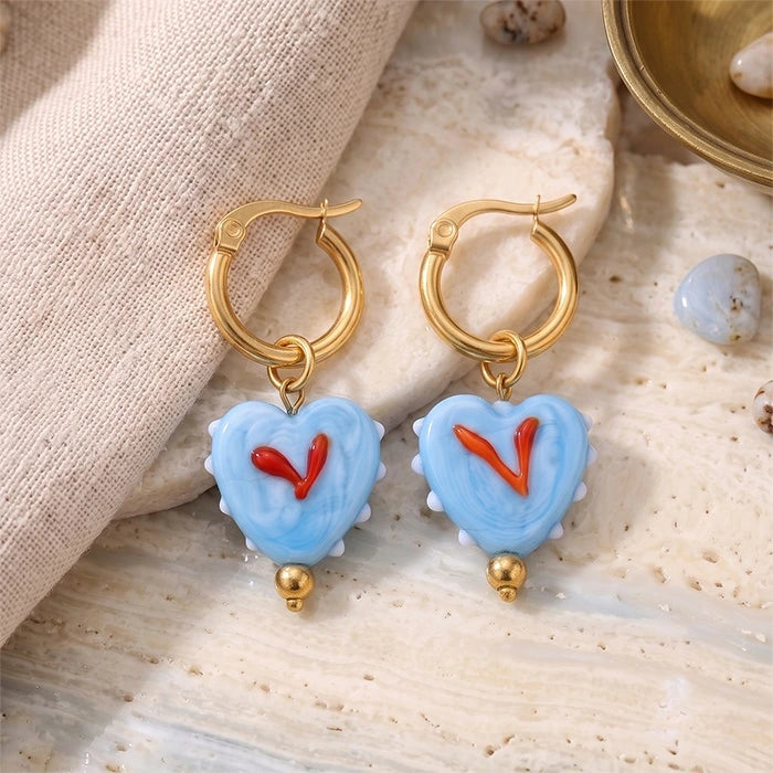 A pair of gold hoop earrings with light blue heart-shaped charms featuring red 'V' initials, displayed on a textured beige stone surface next to a beige fabric napkin.