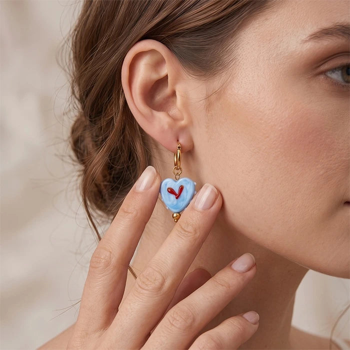 A close-up lifestyle shot of a woman wearing a light blue heart-shaped drop earring with a small red heart in the center and gold-tone metal hardware.