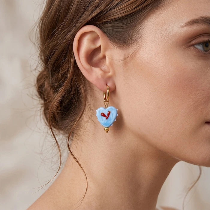 A close-up of a woman's ear wearing a light blue heart-shaped drop earring with a small red heart in the center and gold-tone metal details.