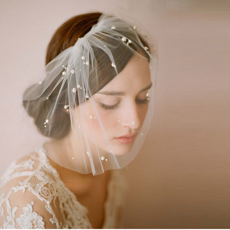 A woman wearing a sheer white bridal veil adorned with small pearl-like beads, styled over her hair. She is wearing a white lace bridal gown against a soft, warm beige background.