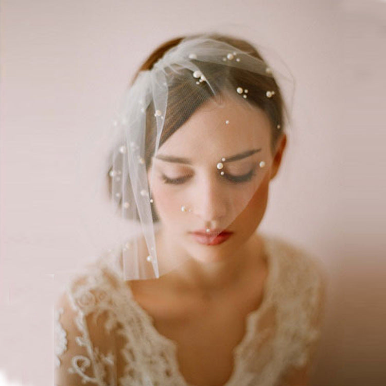 A close-up portrait of a woman wearing a sheer white bridal veil adorned with small pearl-like beads and a cream lace dress against a soft pink background.