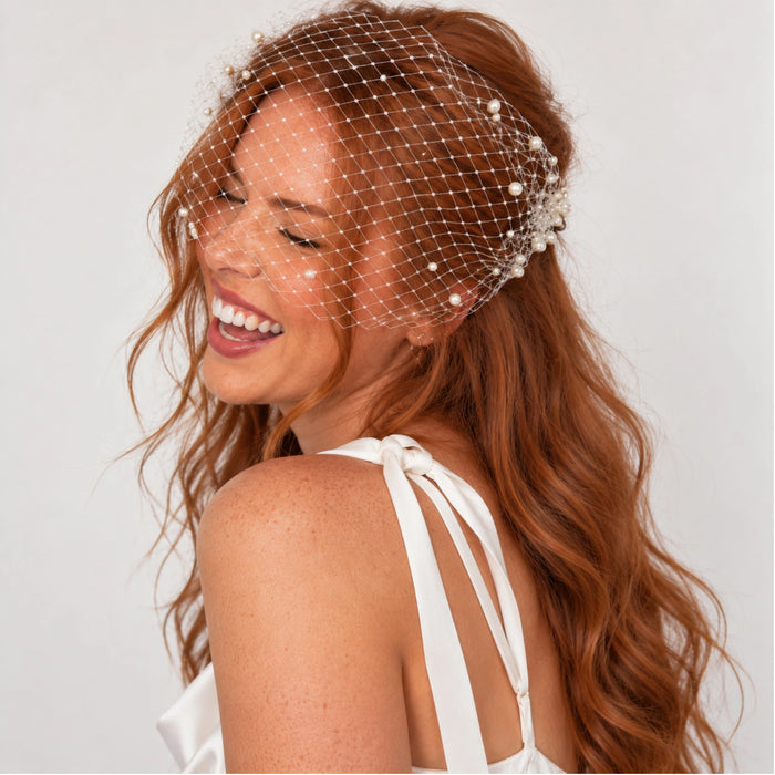 A smiling bride-to-be with long wavy red hair wears a pearl-beaded bridal veil.
