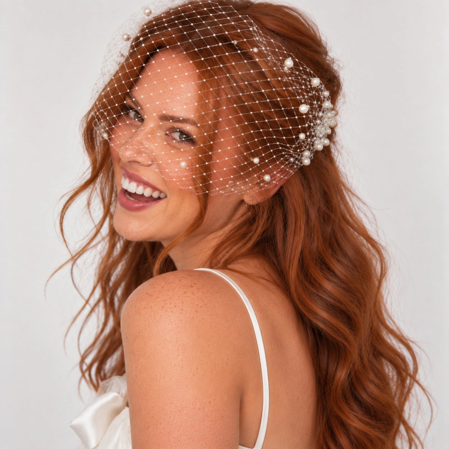 A smiling bride-to-be with long wavy red hair wears a sheer white netted bridal headpiece decorated with small pearls.