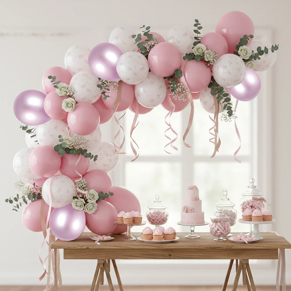 A pink and white balloon arch decorated with green foliage and ribbons, paired with a matching dessert table featuring cupcakes, candy jars, and a small cake in a bright indoor setting.