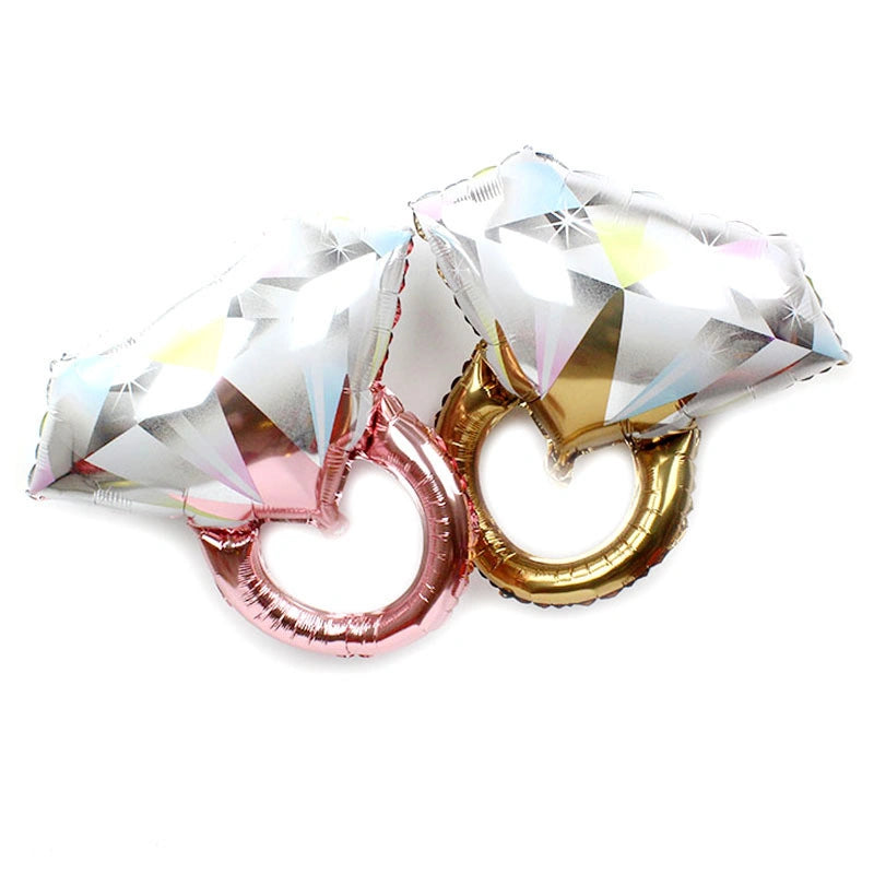 Two metallic foil balloon decorations shaped like oversized diamond rings, one in rose gold and one in gold, with faceted silver diamond tops on a plain white background.