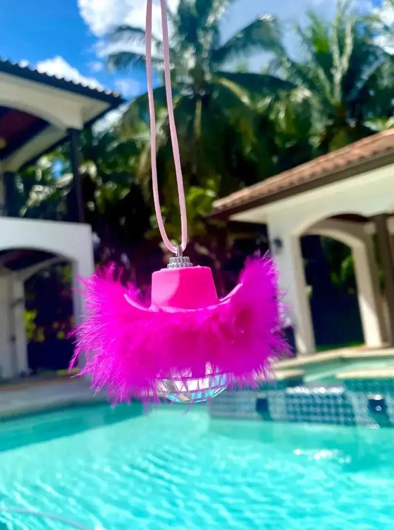 Pink feather disco ball car charm hanging in front of a swimming pool.