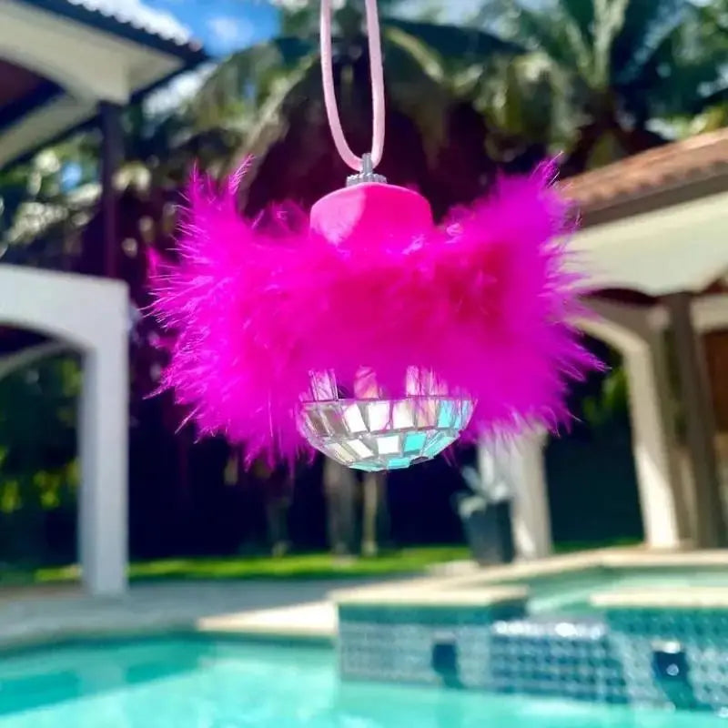 Pink feather disco ball car charm hanging in front of a swimming pool.