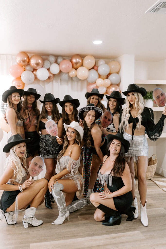 Bride tribe in disco cowgirl outfits posing indoors beneath a blush, white, and rose gold balloon garland at a hen party.