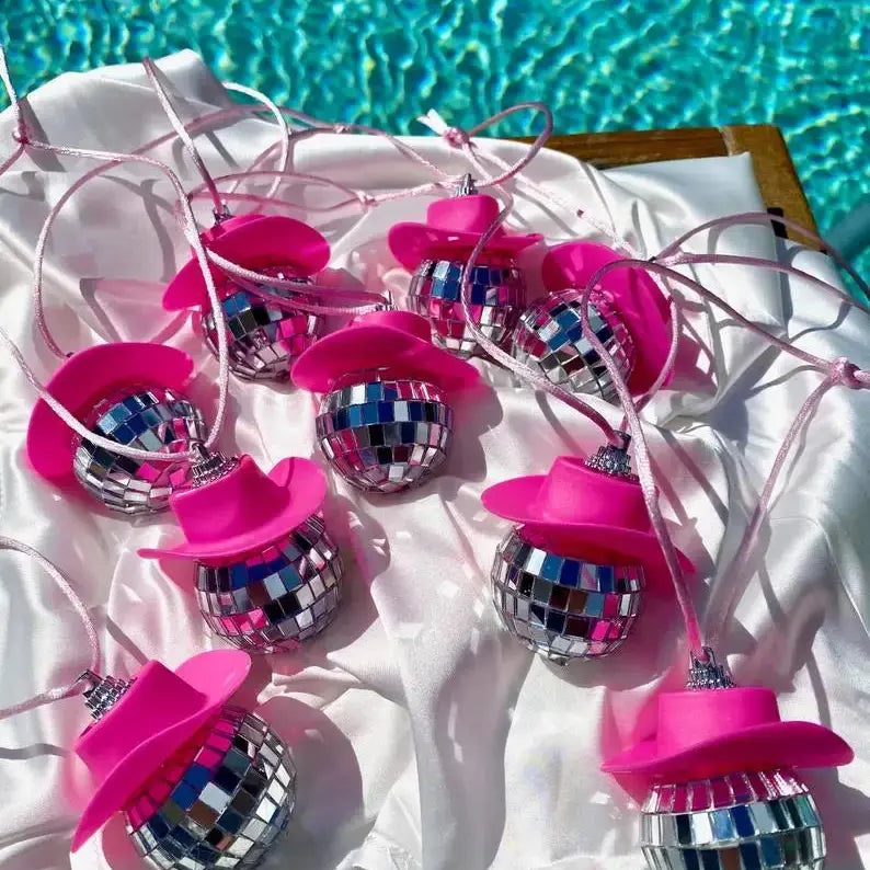 Pink disco cowgirl car charms with mini cowboy hats and mirrored disco balls displayed on white satin beside a swimming pool.