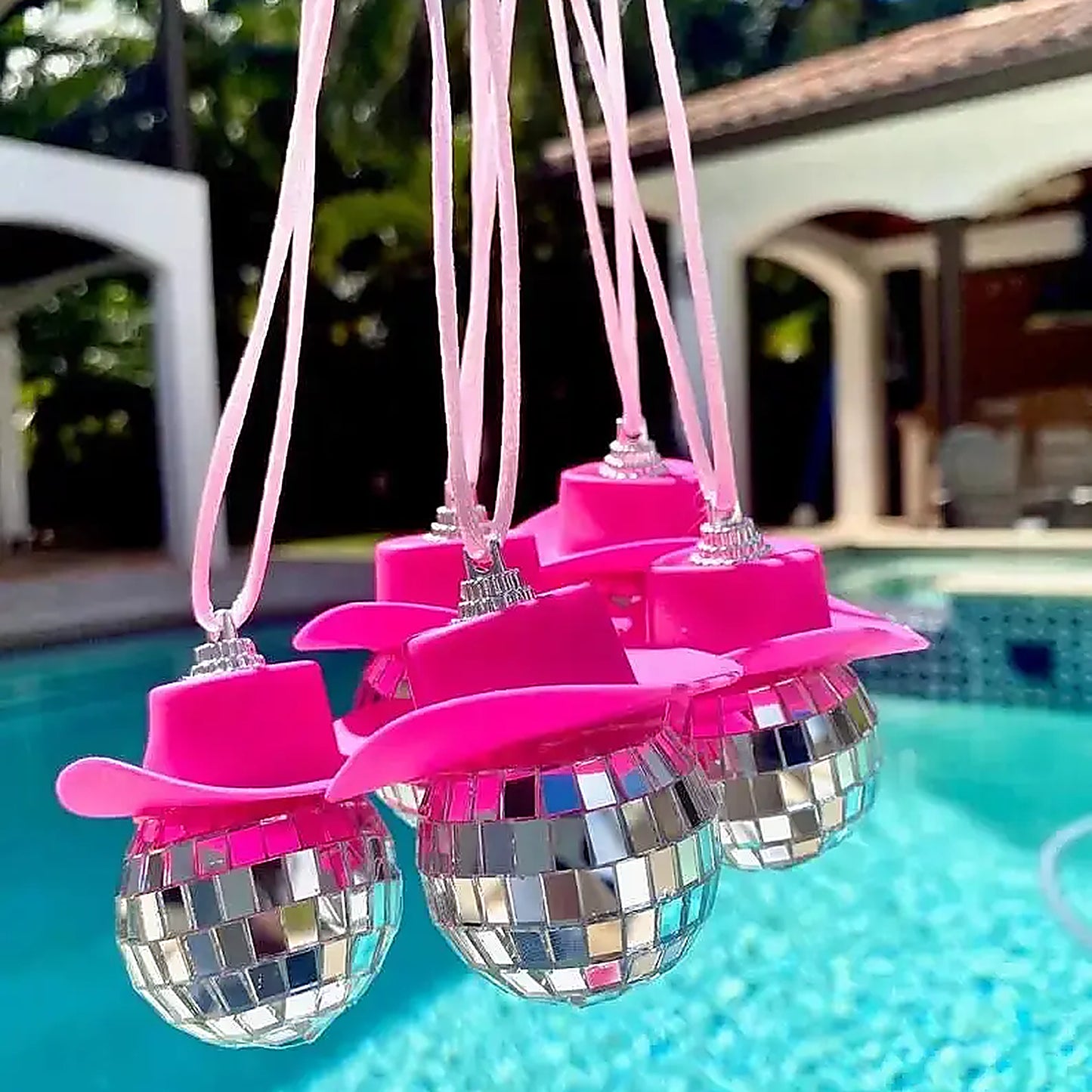 Pink disco cowgirl car charms with mini cowboy hats and mirrored disco balls hanging in front of a swimming pool.