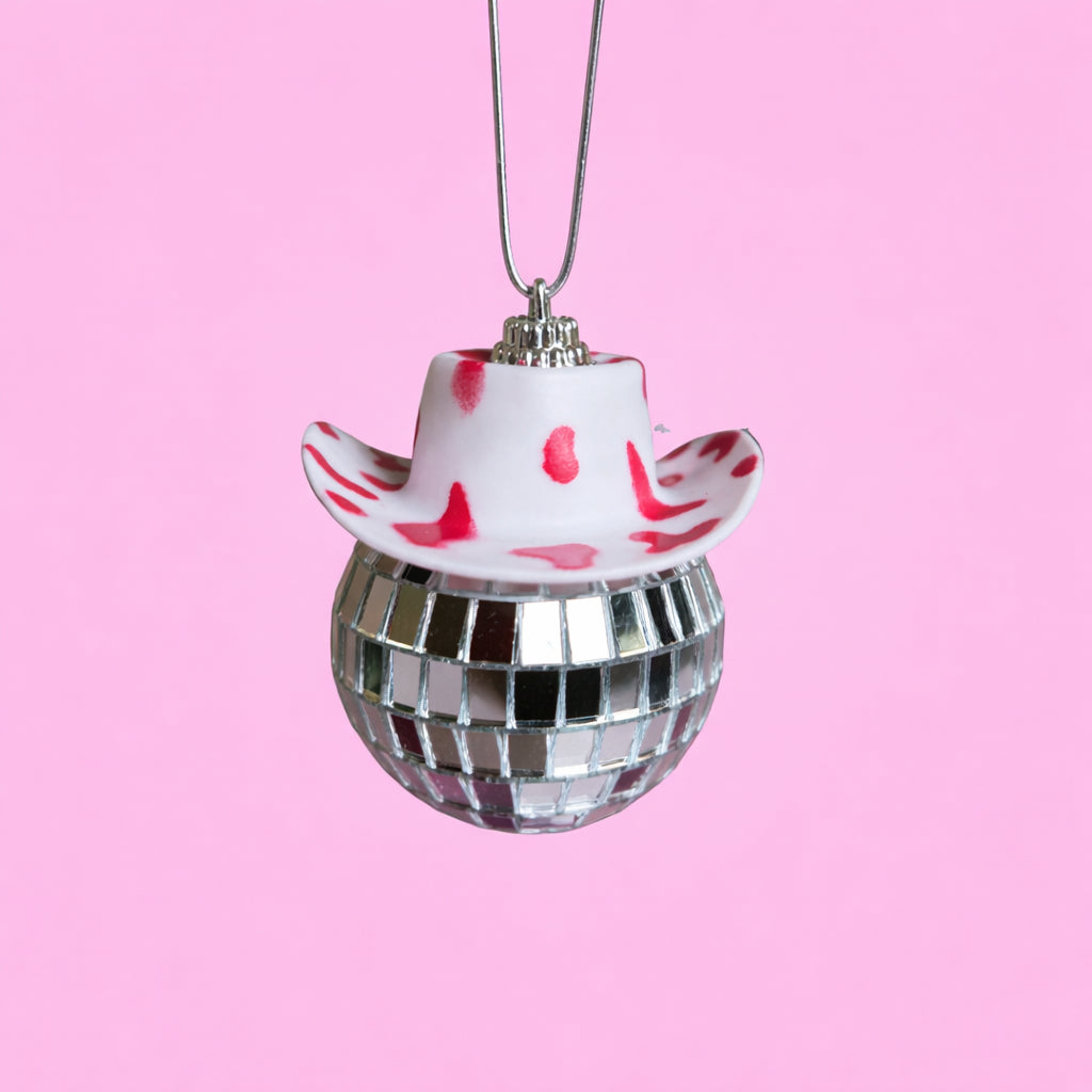 Red and white disco cowgirl car mirror charm with a cow print mini cowboy hat and mirrored disco ball on a soft pink background. 