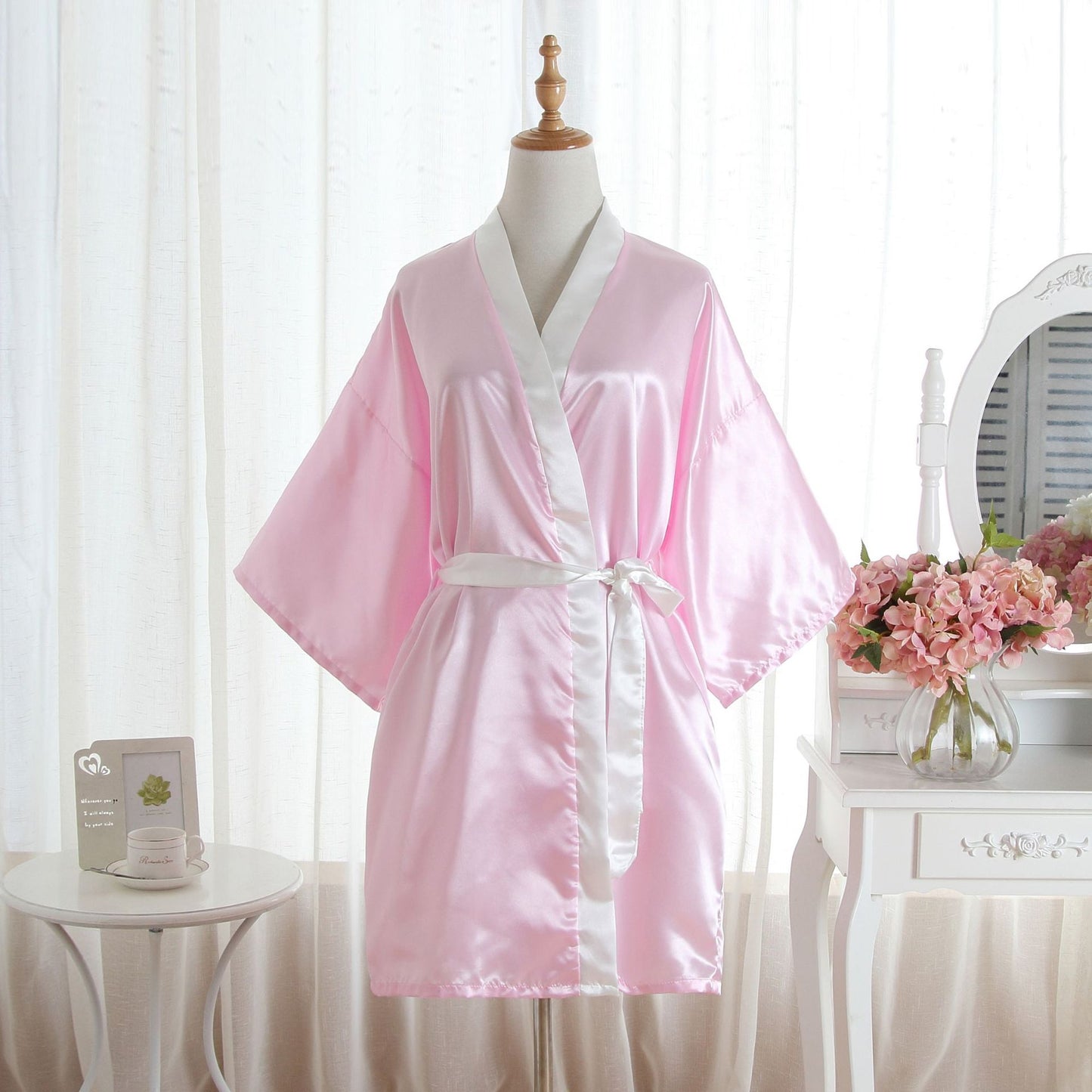 A light pink satin robe with white trim and a white tie belt, displayed on a mannequin in a softly lit room with white curtains.