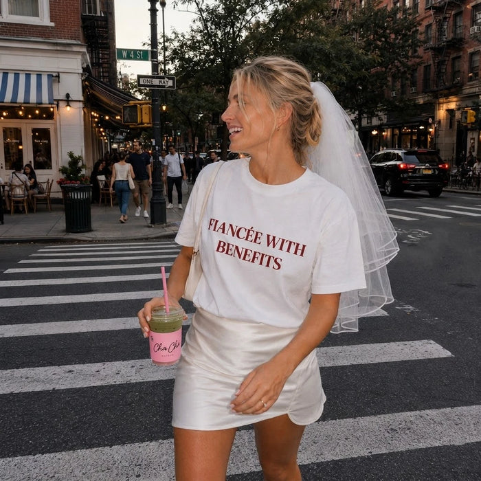 Woman in a white t-shirt with 'FIANCEE WITH BENEFITS' text, white skirt, and veil on a city street.