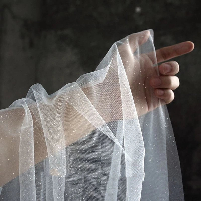 A close-up of a hand holding up a sheer white tulle fabric with tiny scattered glitter or speckle details against a dark textured background.