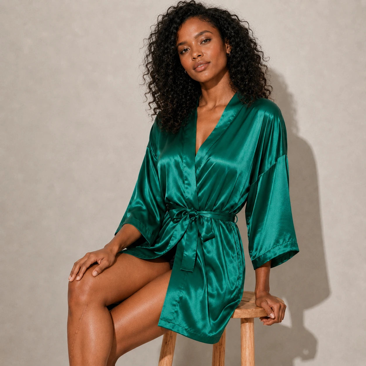 Woman wearing a green satin robe sitting on a wooden stool against a beige background