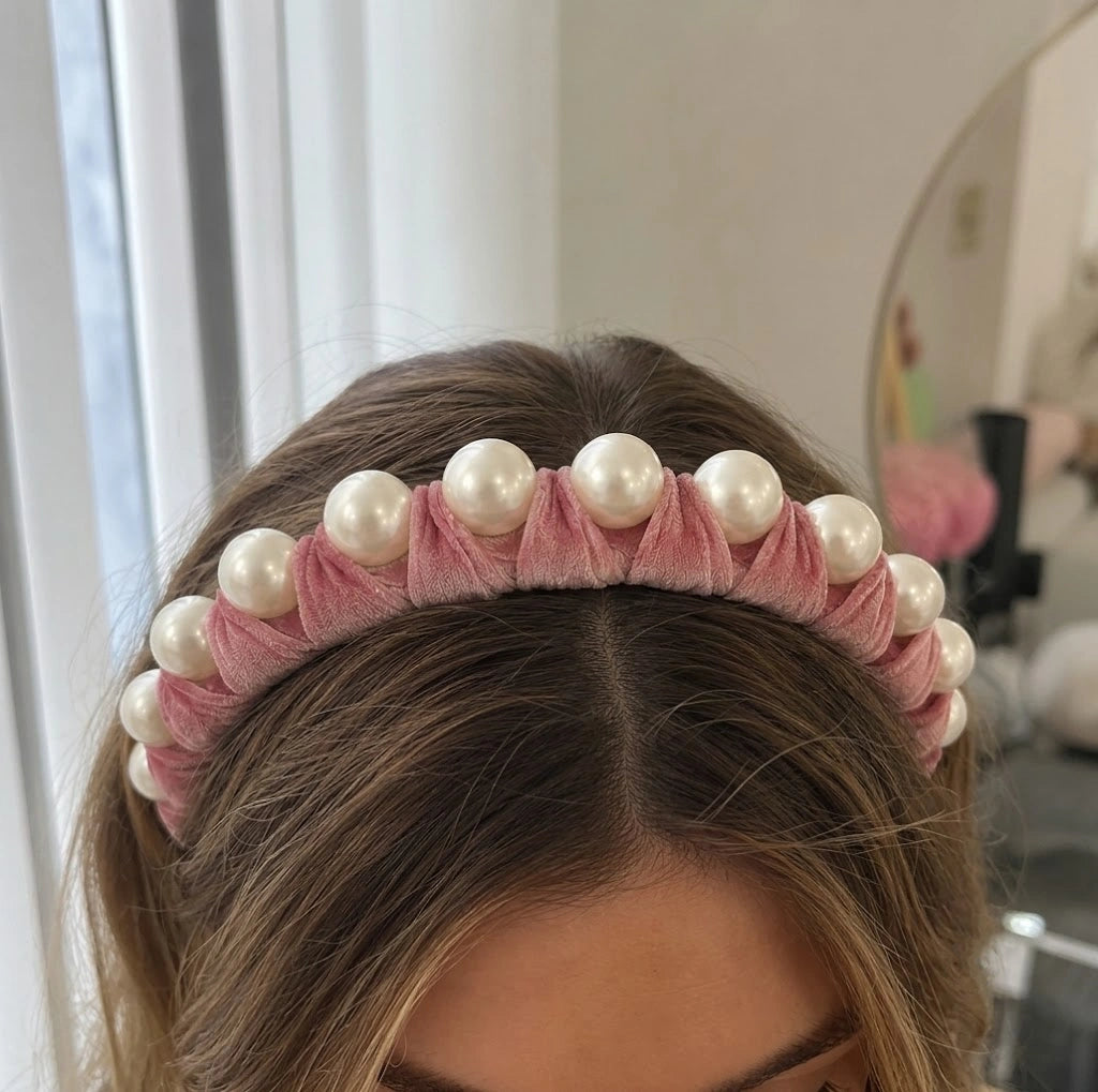 A pink velvet headband with a row of white faux pearls, worn by a model with brown hair.