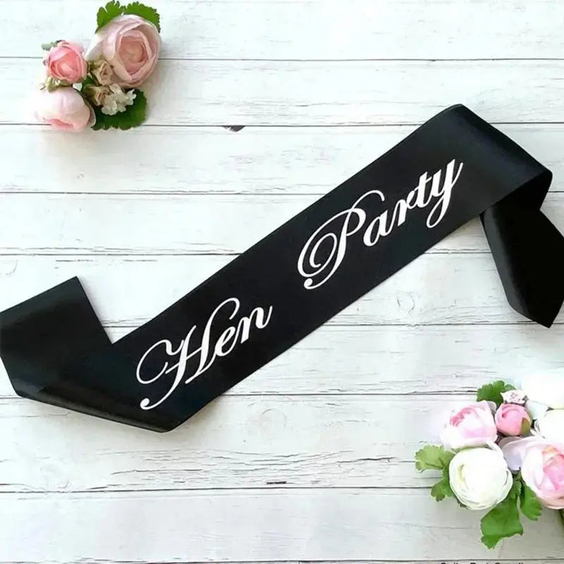 Black sash with 'Hen Party' text on a white wooden background with flowers.