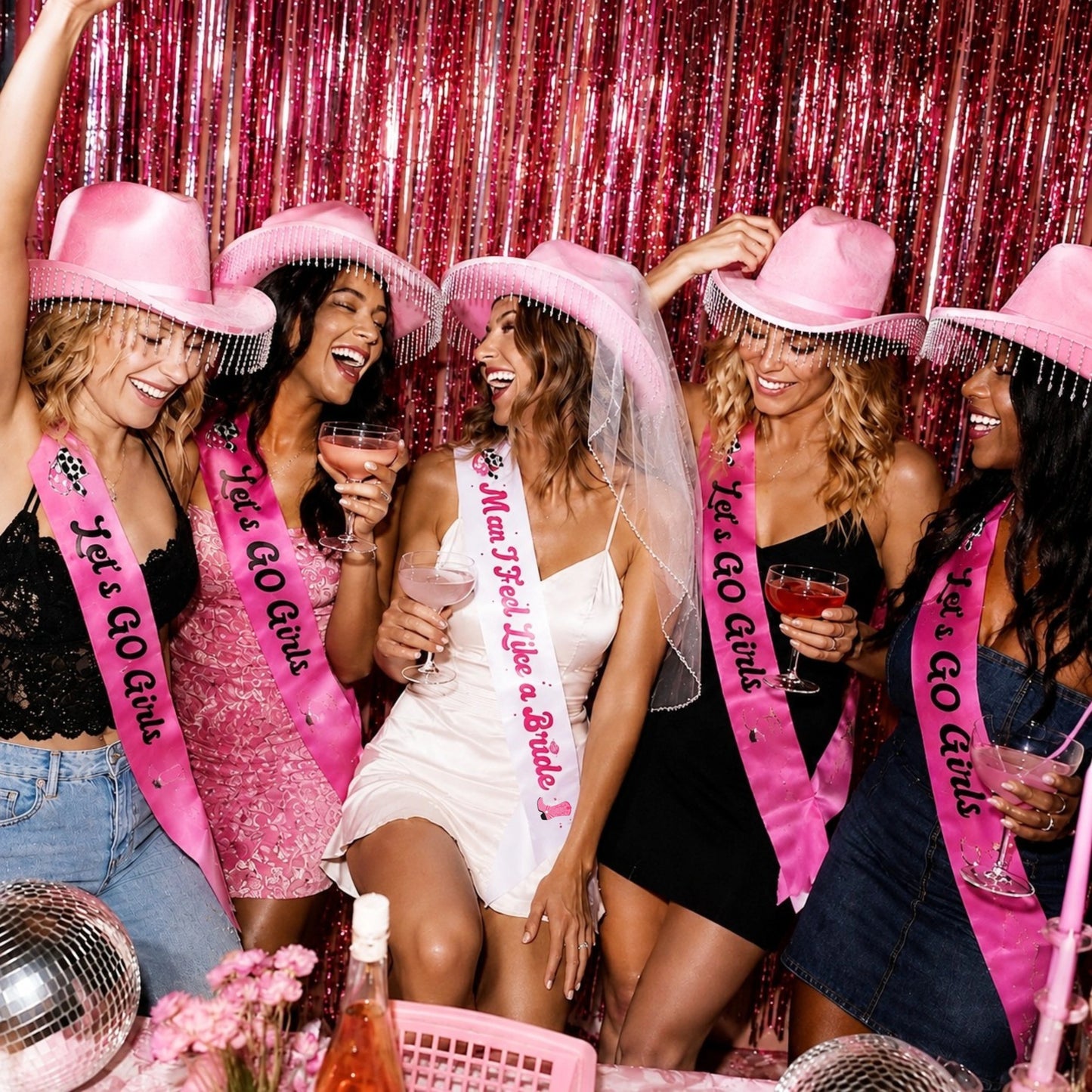 Five women celebrating at a party wearing pink glittery cowboy hats, pink sashes that read 'Let's Go Girls,' and holding pink drinks in front of a pink tinsel backdrop.