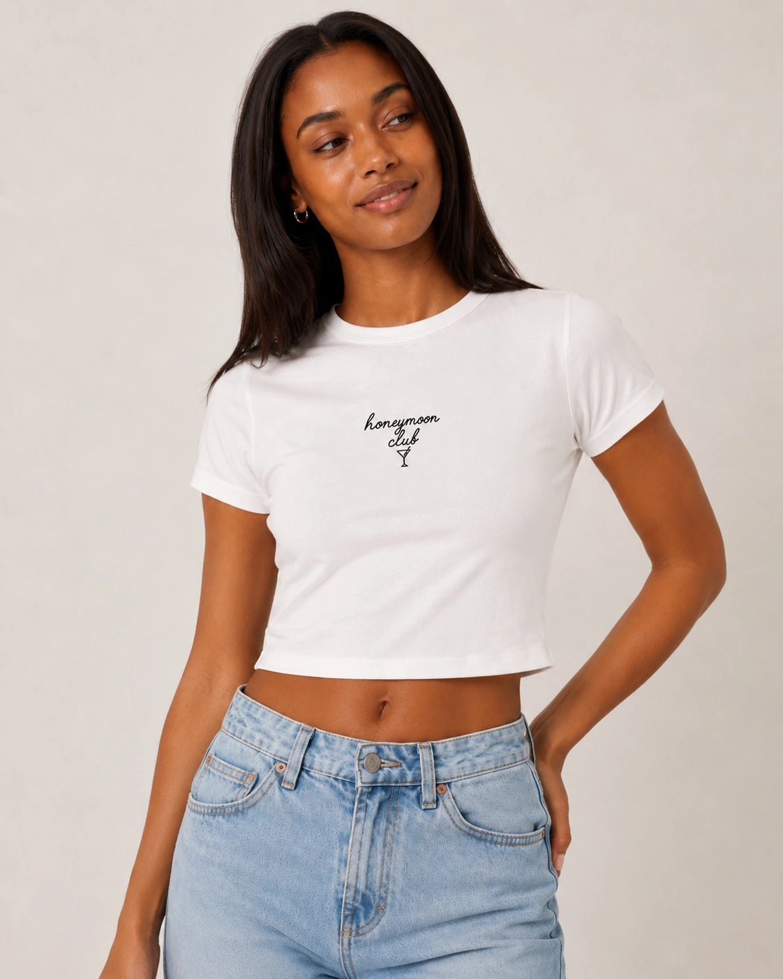 Woman wearing a white t-shirt with 'honeymoon club' text and light blue jeans on a plain background