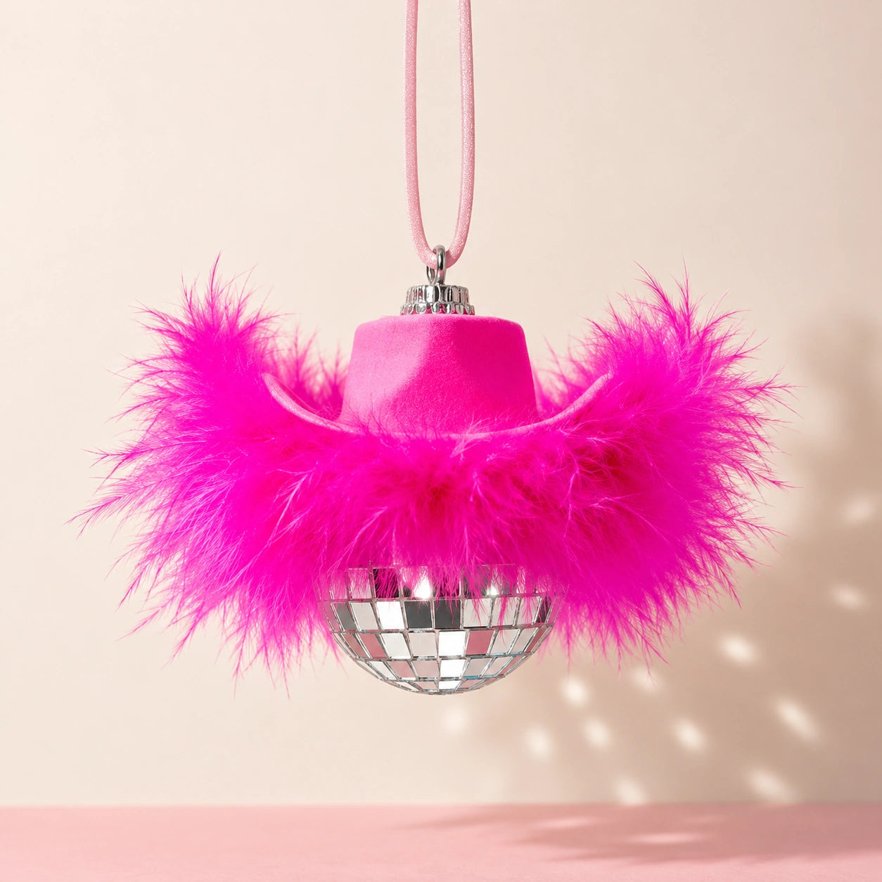A pink cowboy hat ornament with bright hot pink feathers and a mirrored disco ball underneath, hanging by a pink ribbon against a warm beige background.