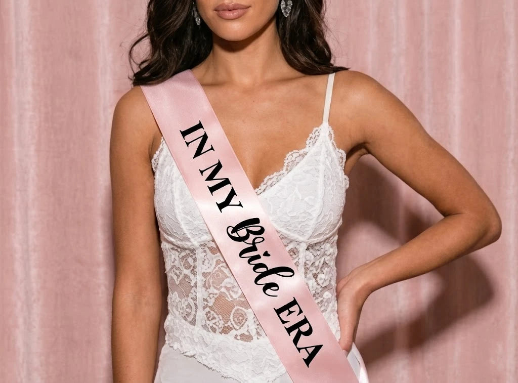 Woman wearing a white lace dress with a pink sash that reads 'IN MY Bride ERA' against a pink background.
