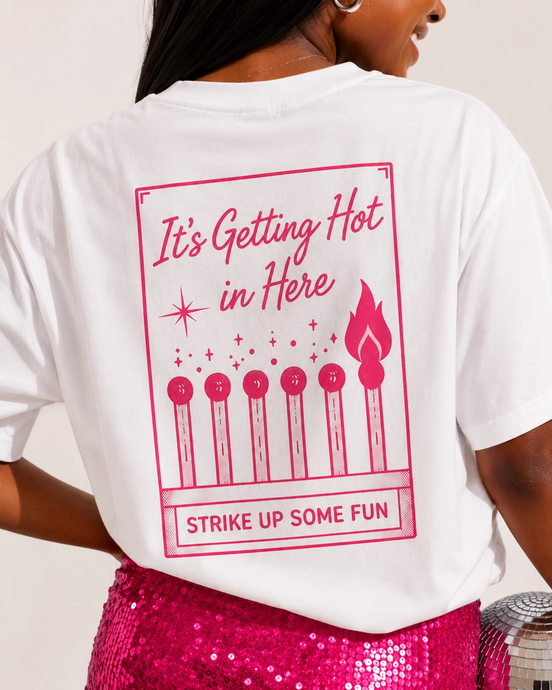 A woman shown from the back wearing a white graphic T-shirt with a pink matchstick design and the text 'It's Getting Hot in Here' and 'Strike Up Some Fun.'.