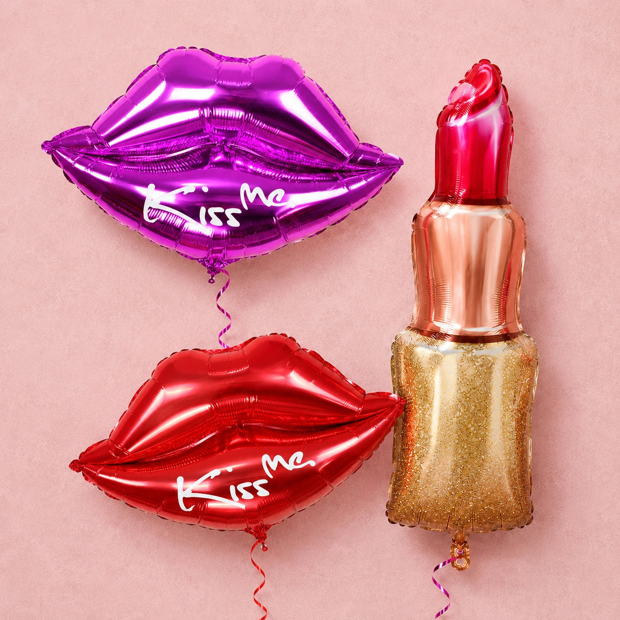 Purple and red Kiss Me lip-shaped balloons beside a gold lipstick balloon on a soft pink textured background.