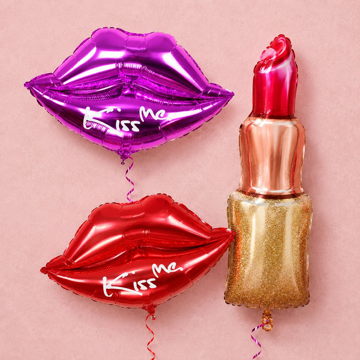Purple and red Kiss Me lip-shaped balloons beside a gold lipstick balloon on a soft pink textured background.