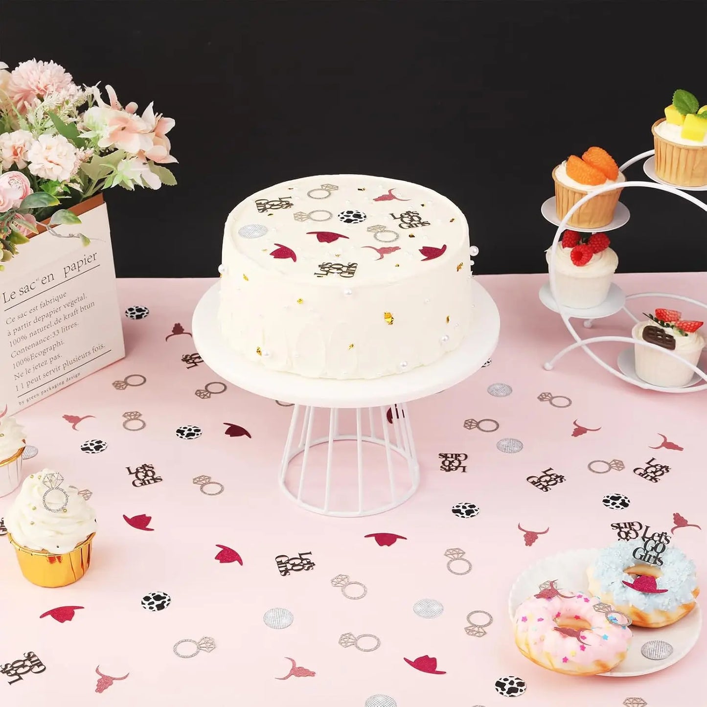 Decorative cake with scattered confetti on a stand with cupcakes and flowers on a pink tablecloth.