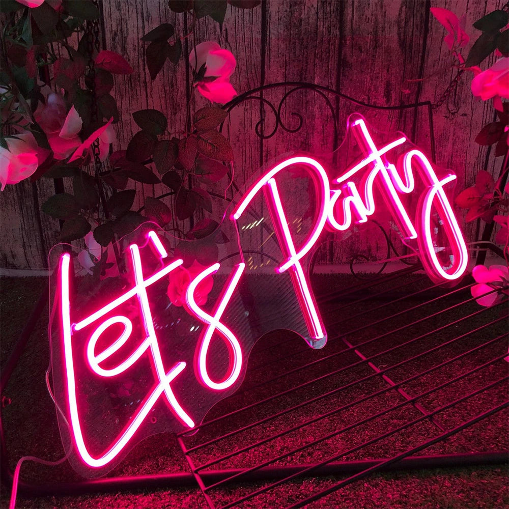 A glowing pink neon sign that reads “Let’s Party” in cursive script, displayed on a metal rack against a rustic wooden wall with artificial flowers.