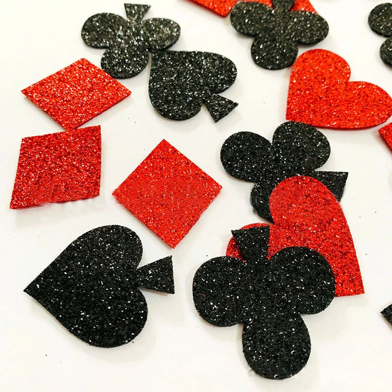 Glittery playing card suit confetti pieces including red and black hearts, diamonds, clubs, and spades scattered on a white background.