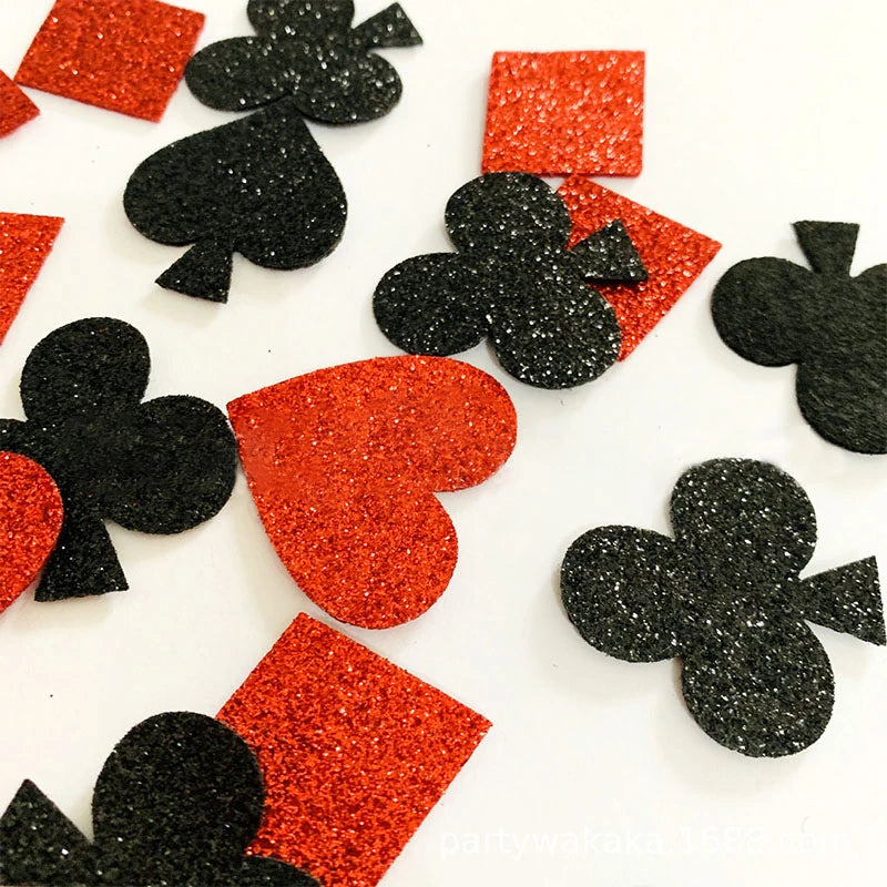 Black and red glitter playing card suit confetti pieces scattered on a white background, including hearts, diamonds, clubs, and spades.