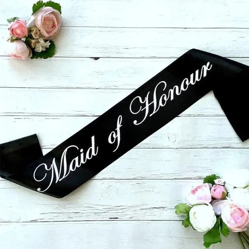 Black sash with 'Maid of Honour' text on a white wooden surface with floral decorations.