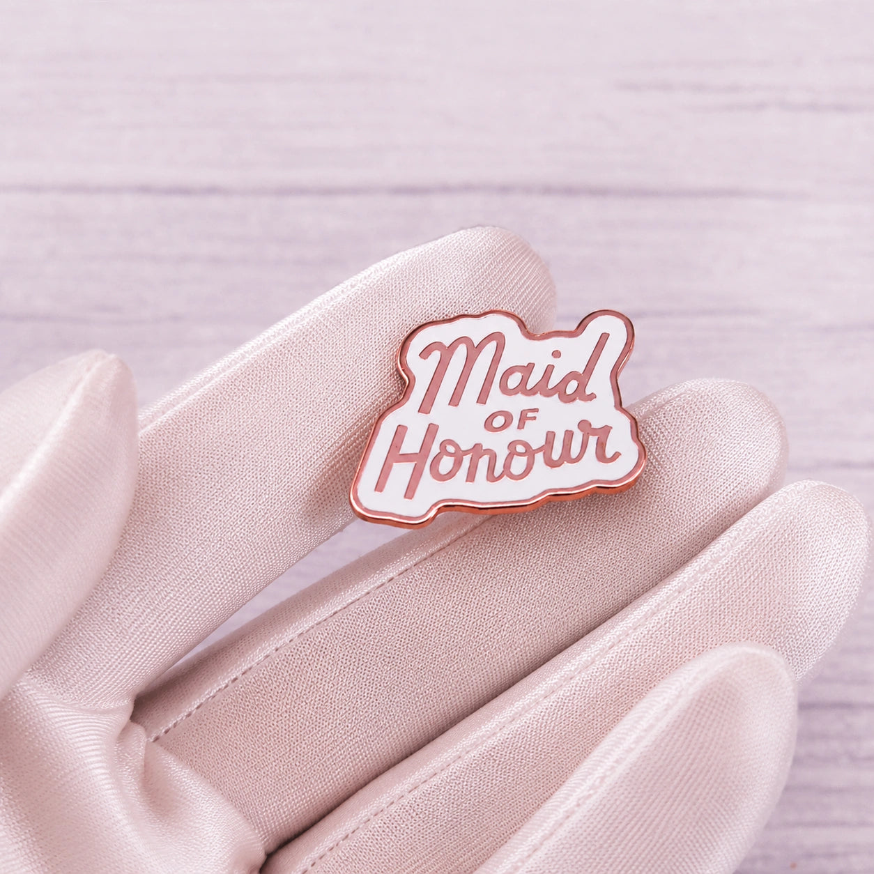 A small enamel pin reading 'Maid of Honour' in pink script on a white background with a rose-gold outline, held on a pale pink satin glove over a light wood surface.