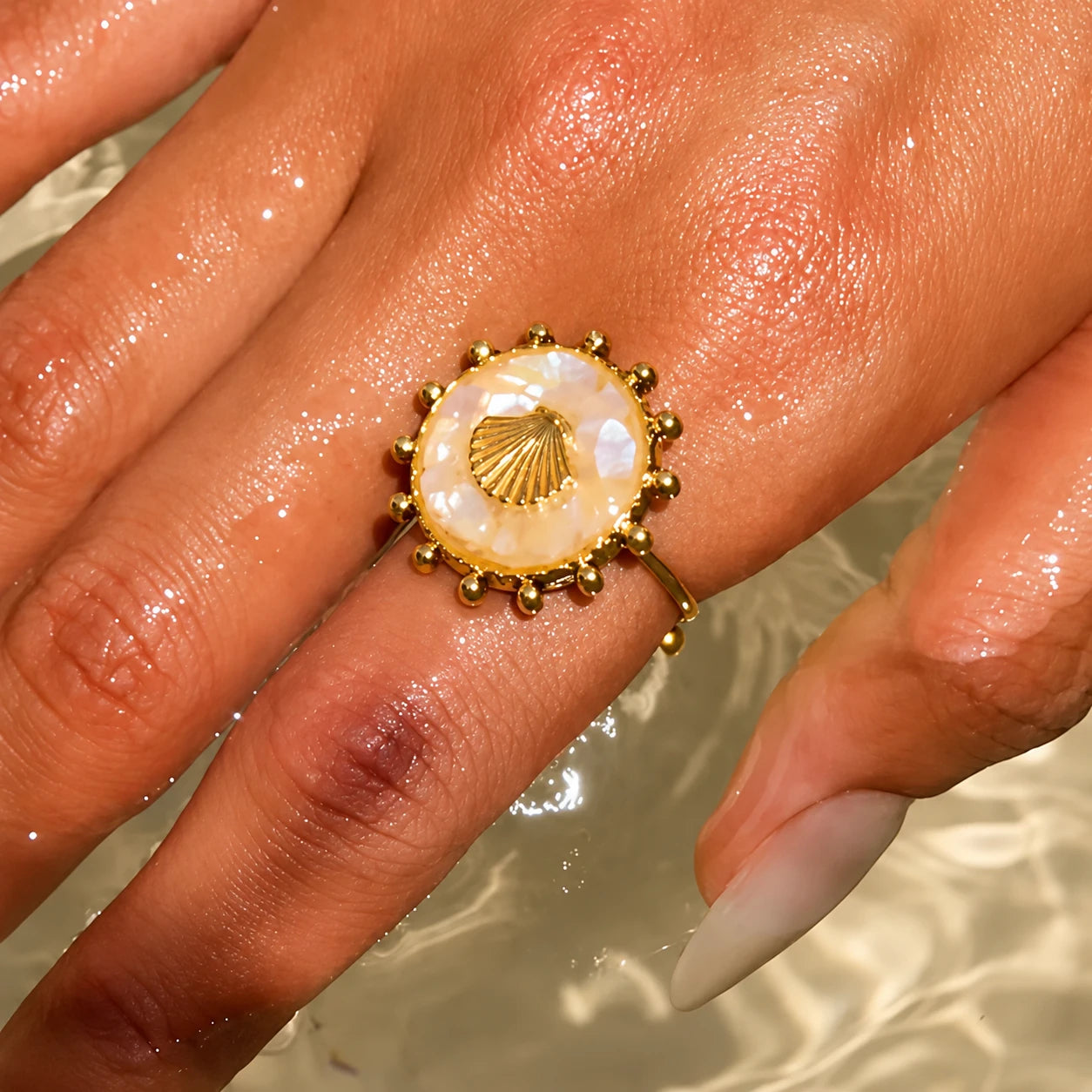 A gold ring with a central iridescent shell-shaped stone and beaded border, worn on a person's finger in shallow water.