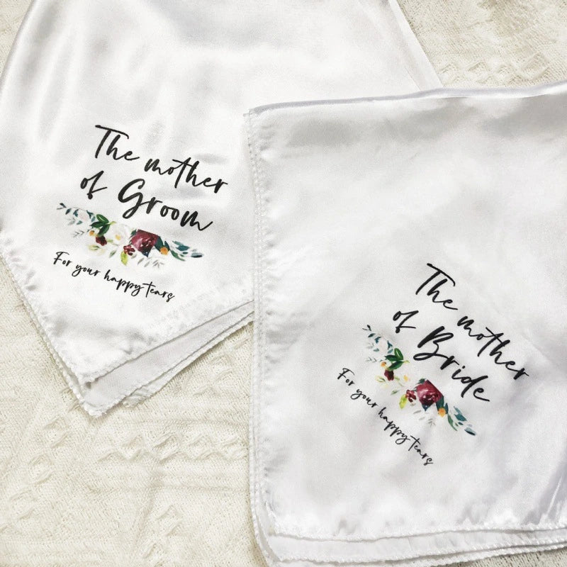 Two white square baby or bridal scarves laid flat on a textured cream fabric background. One scarf reads 'The mother of Groom' with a small floral illustration, and the other reads 'The mother of Bride' with a matching floral illustration.
