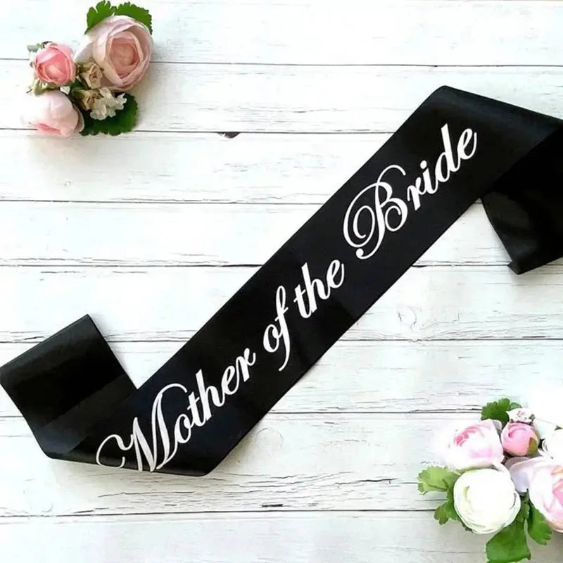 Black sash with 'Mother of the Bride' text on a white wooden background with flowers.